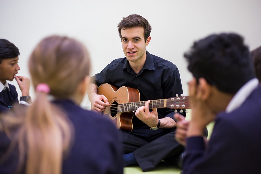 A music teacher is seated on the floor, engaging with a small group of students in a classroom setting. He is holding and playing a guitar, looking at the students with a smile, indicating a warm and interactive learning environment. The students, wearing school uniforms, are attentively watching and listening, suggesting a focus on music education and the enjoyment of learning an instrument together.