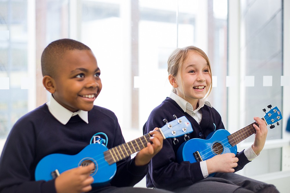 Two joyful schoolchildren are seated side by side, each holding a bright blue ukulele. They are engaged in a music lesson, with the boy looking to his side with a bright smile, and the girl looking ahead, laughing as she strums her instrument. The natural light from the window behind them highlights the cheerful atmosphere of their learning experience.
