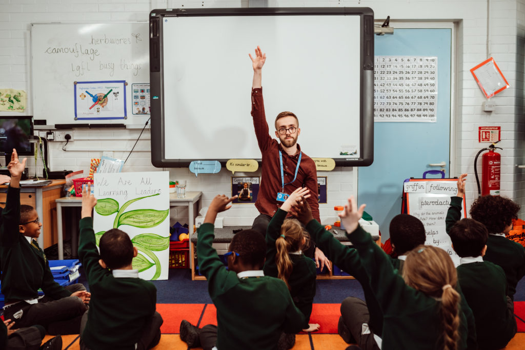 Male sitting at the front, ten primary pupils sitting in front of him, male and pupils hands up in air.