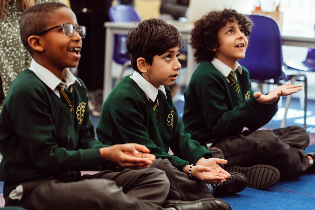 Three schoolchildren in green sweaters sit cross-legged on the floor, their expressions ranging from joyful to attentive as they participate in a classroom activity. The child on the left is smiling broadly, the middle one looks on with focused interest, and the one on the right is animatedly engaged, all embodying a lively and interactive learning environment.
