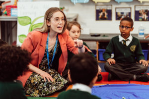 A vibrant classroom scene unfolds with a teacher in a coral blazer and floral dress, animatedly engaging with her students seated on the floor. Her expression is enthusiastic as she points, capturing the attention of the children in school uniforms who listen intently, embodying a moment of interactive learning. The backdrop of educational posters and student work adds to the atmosphere of active and joyful education.