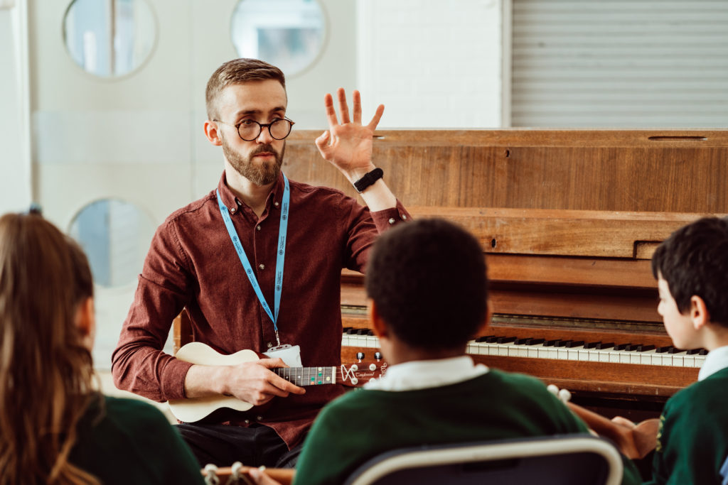 A music teacher with a beard and glasses is holding a ukulele and gesturing with his hand, engaging with young students in a classroom setting. The students, wearing green school uniforms, are attentively listening to the teacher. A piano is visible in the background, suggesting a comprehensive musical learning environment. The atmosphere is one of focused instruction and interactive learning.