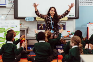 A joyful teacher with outstretched arms leads a lively classroom activity, surrounded by engaged young students in green uniforms, all sitting on the floor and participating with raised hands. The classroom is adorned with educational materials and colorful displays, creating an atmosphere of interactive learning.