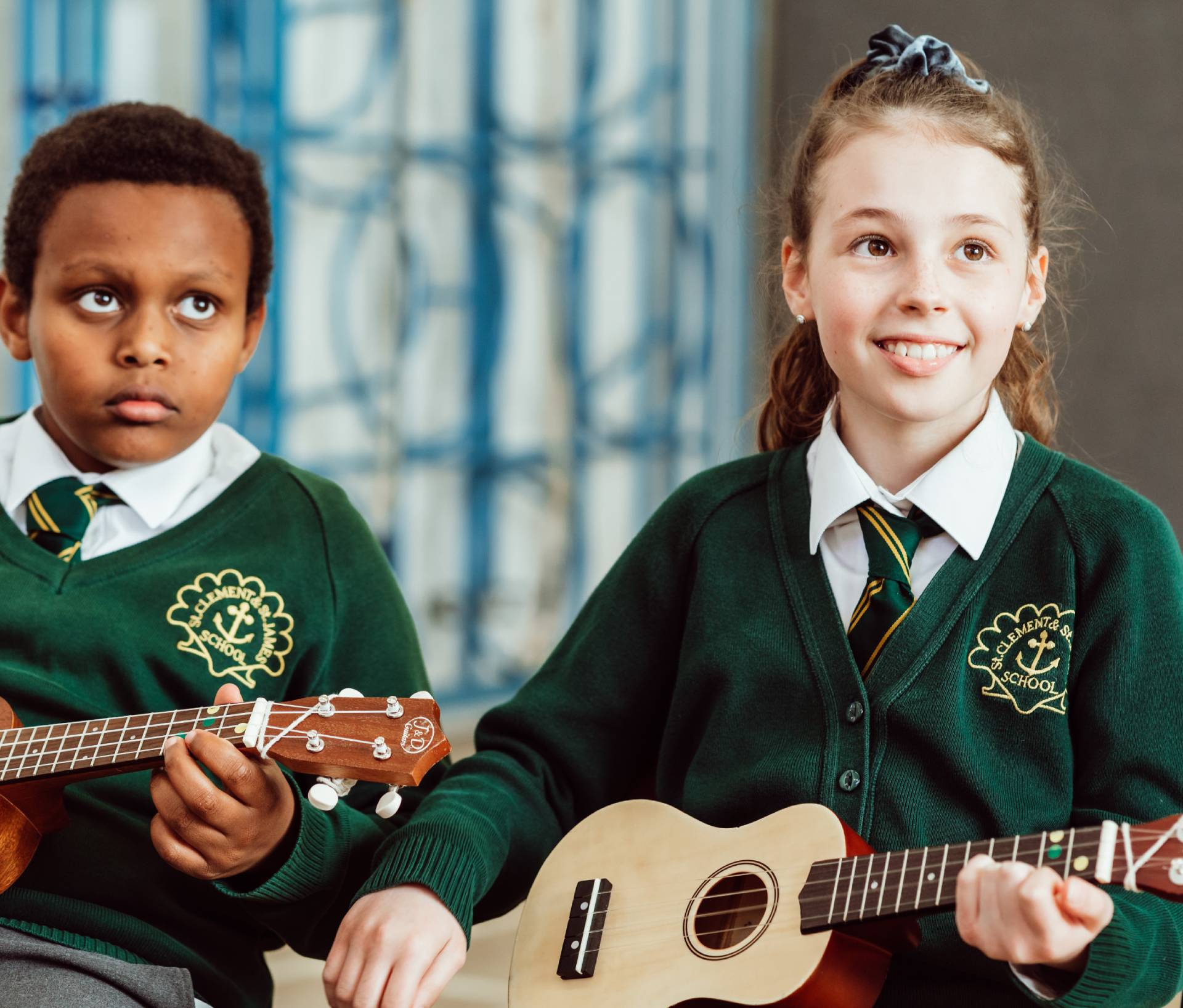 Two KS2 primary pupils, holding Ukuleles, sitting in school hall, on chairs, both pupils holding brown Ukuleles, smiling.