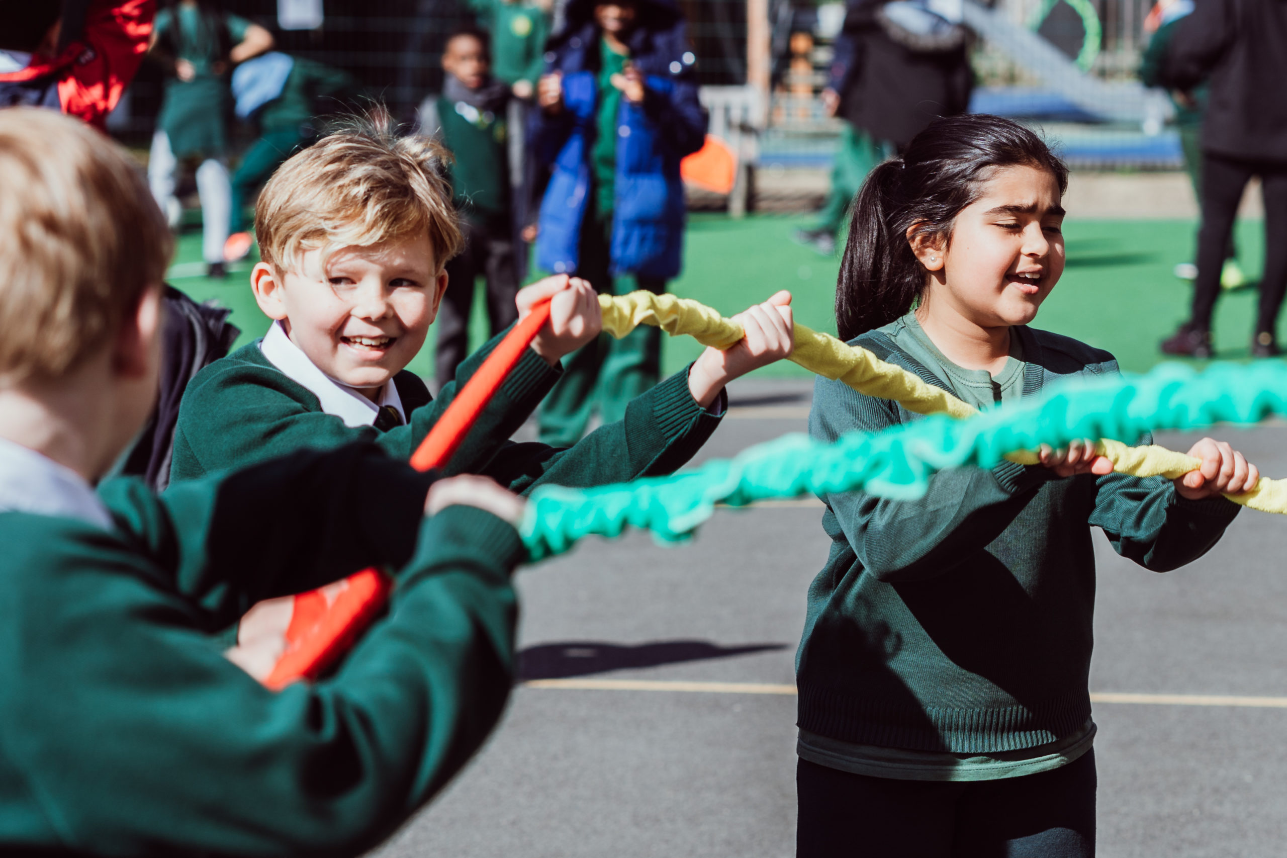 Three KS2 primary pupils holding a red, yellow, green giant scrunchie singing in the playground.