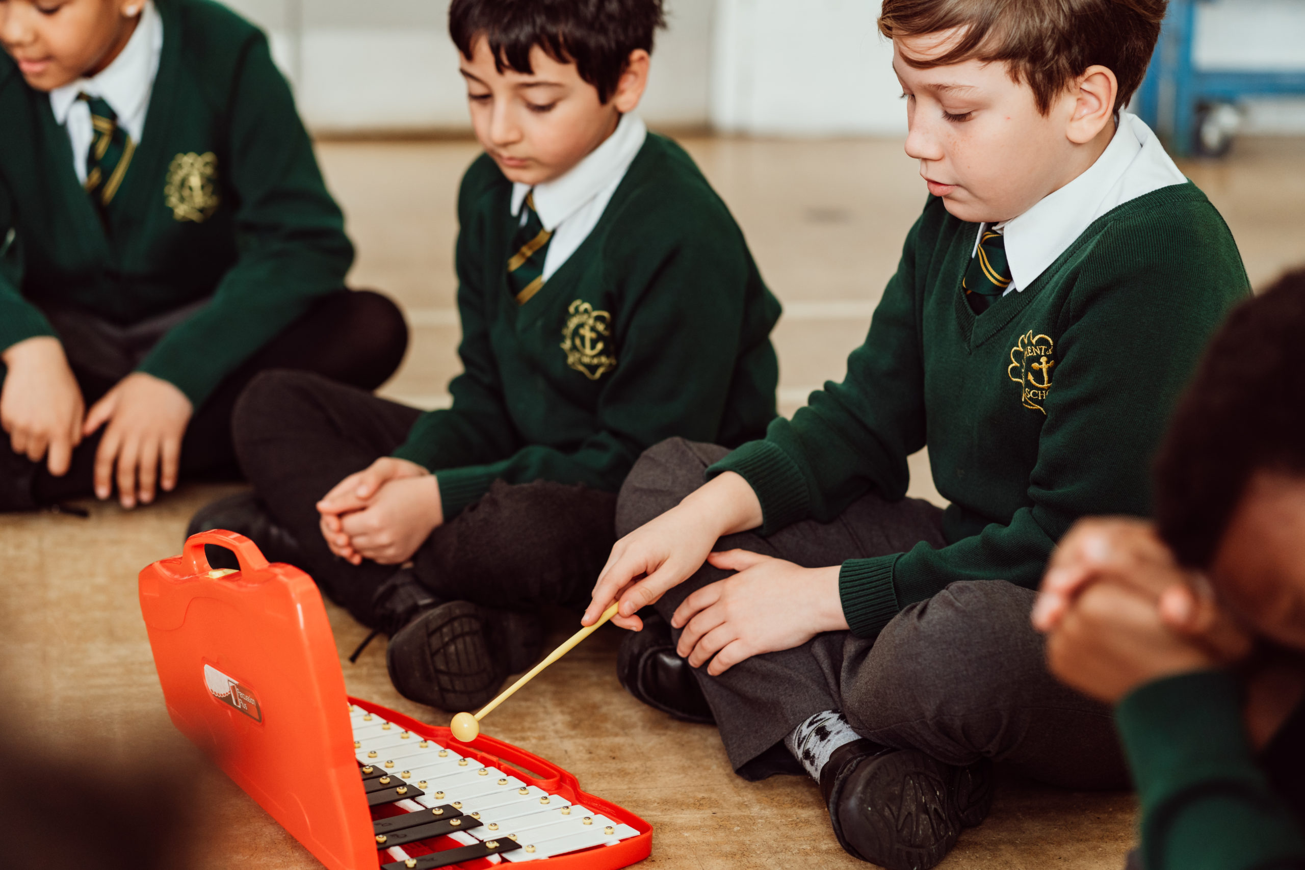 Three KS2 primary pupils sitting on floor of hall, red xylophone in front of them, one pupil holding yellow beater