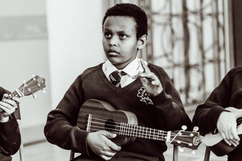 Close up- black and white, KS2 primary pupil, holding ukulele in one hand, holding his other hand up in air