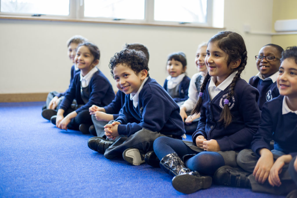 Ten KS1 primary pupils sitting on classroom carpet, legs crossed, in lines, smiling, blue and grey uniform, blue carpet