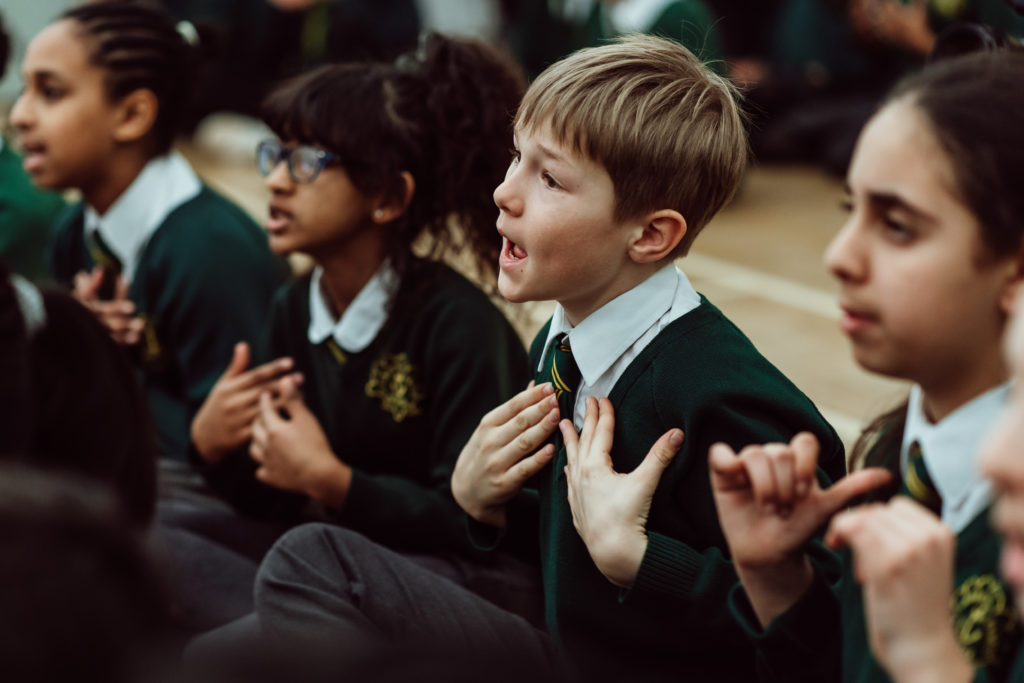 Four KS2 primary children sitting on assembly floor, singing a song. three female pupils and one male, using hand gestures