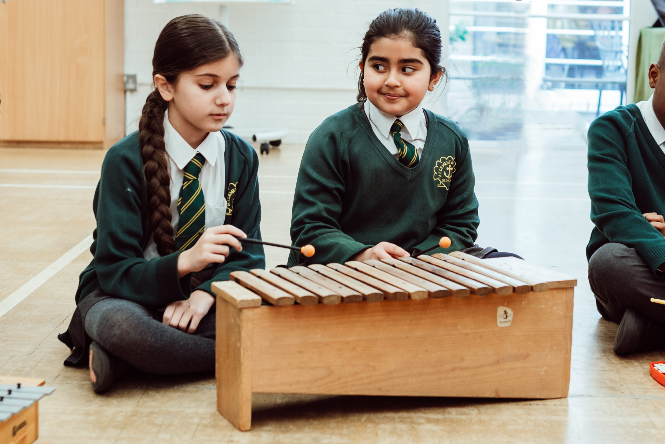 Two schoolchildren in green uniforms are seated on the floor, sharing a wooden xylophone. The girl on the left, with a long braid, holds a mallet and appears focused on the instrument, while the girl on the right looks at her with a smile, suggesting a moment of musical collaboration and learning.