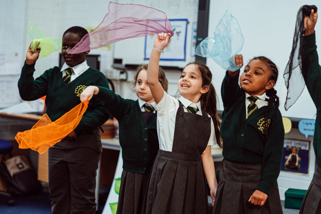Five KS1 primary pupils, standing, waving a blue, pink, orange, black, yellow scarves in the air, smiling.