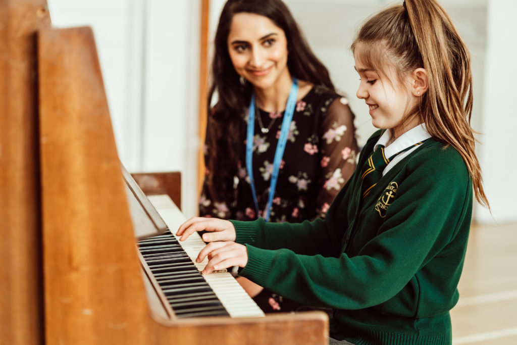 Female and KS2 primary pupil sitting at the piano, pupil playing piano, female watching in the background with a smile.