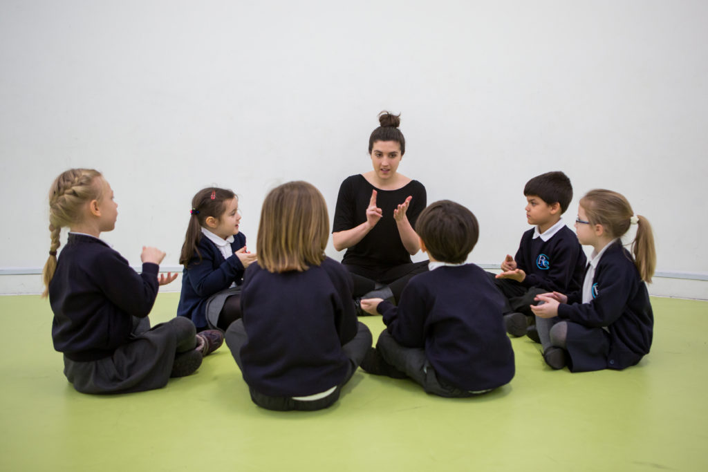 Female sitting on floor, in circle with six KS1 primary pupils, all using both hands for music activity