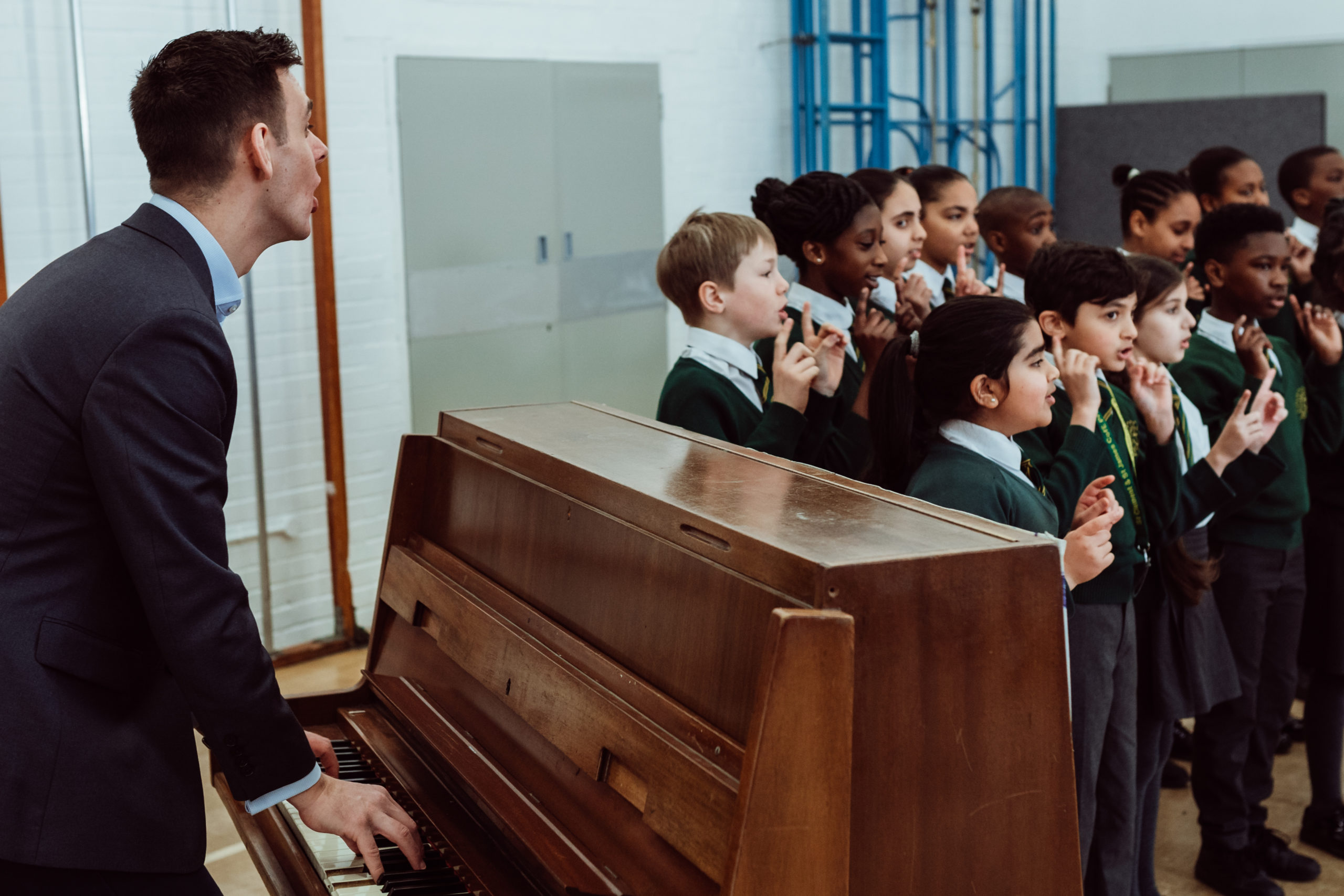 Male playing piano, singing with a choir of primary children, standing in 2 lines, teacher to the left of pupils.