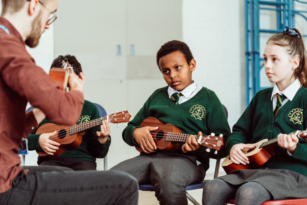 Male and three primary KS2 pupils, all holding ukuleles, all sitting on chairs in the hall.