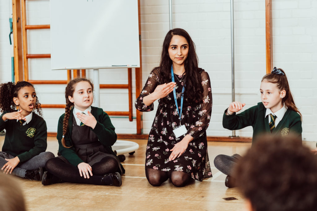 Female kneeling on the floor with hand out in front of her, three primary pupils sitting next to her copying.