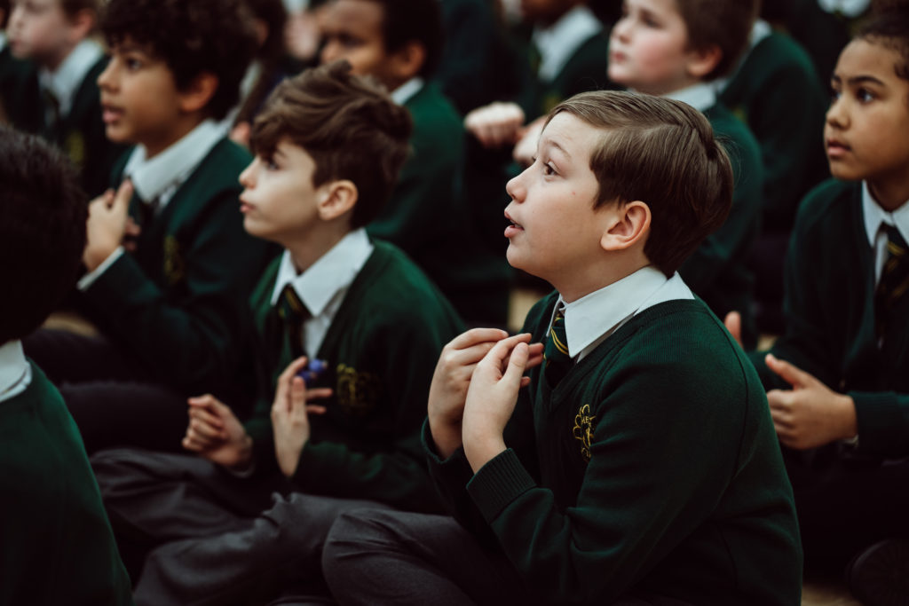 Primary pupil, boy sitting on the floor, holding hands up to his chest, looking forward, six pupils in the background.