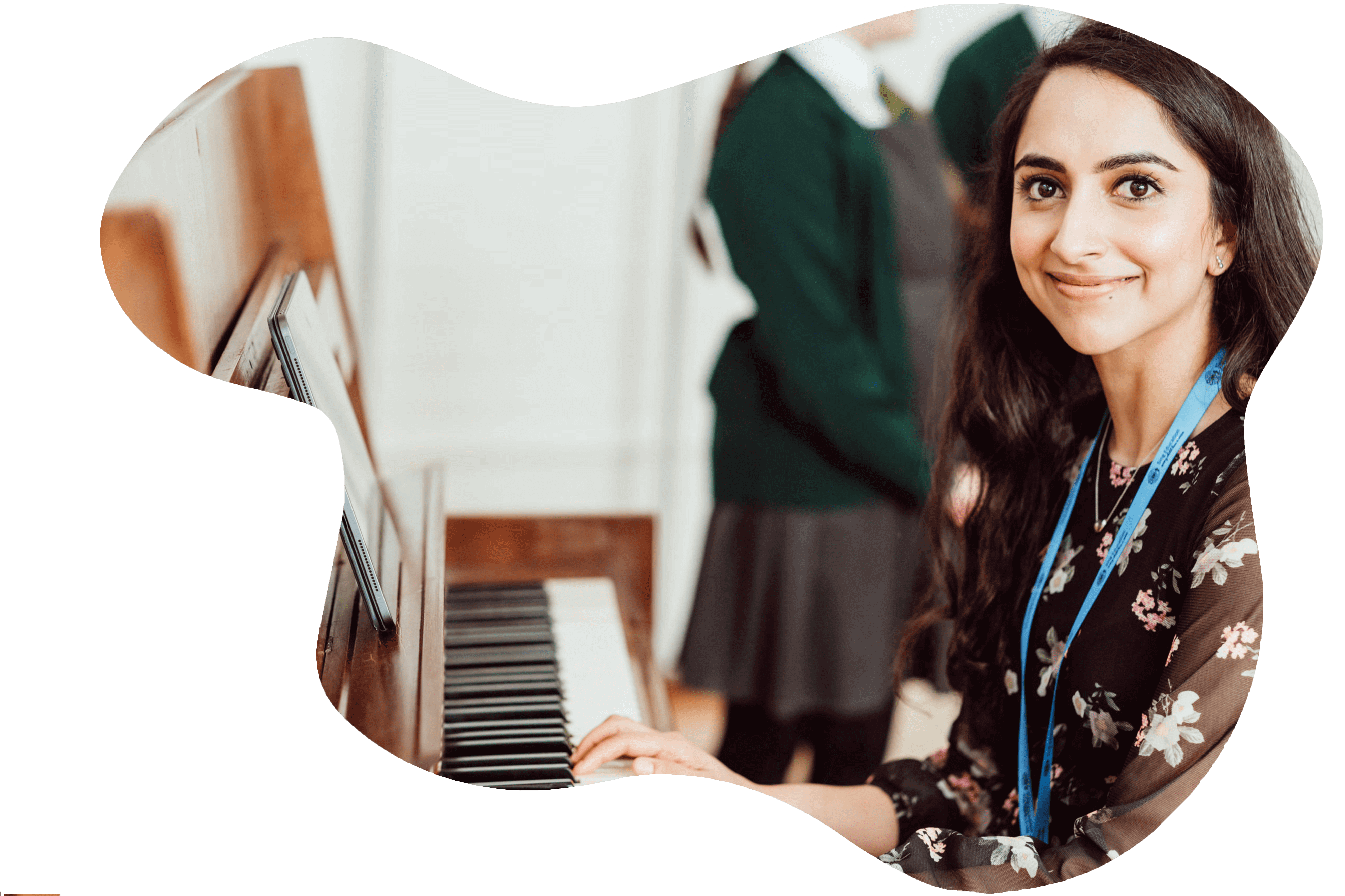 A smiling woman with a lanyard sits at a piano, her hands poised on the keys, ready to play. Behind her, the blurred figure of a student in a green school uniform suggests a classroom setting, emphasizing the educational and musical context of the scene. The woman's engaging expression and the presence of the piano highlight the interactive and creative nature of the learning environment.