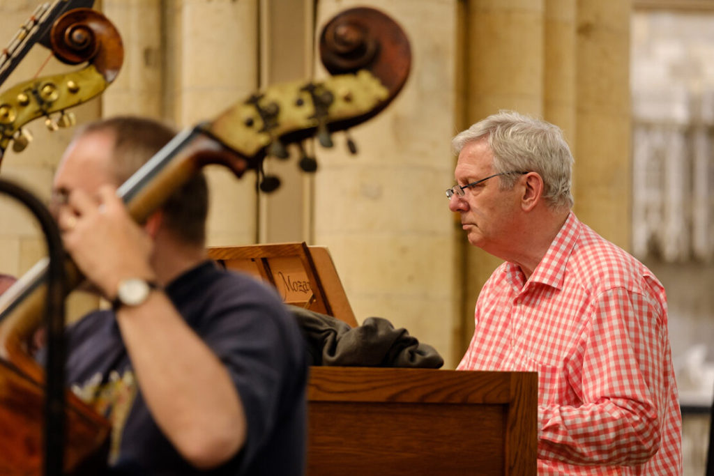 The photo captures a moment during a classical music rehearsal, focusing on a man in a red checkered shirt who appears to be a conductor or musician, intently observing a score or performance. In the foreground, out-of-focus double basses with their curved scrolls add a sense of depth and context to the scene, suggesting an orchestral setting. The warm lighting and the concentration on the man's face convey a serious dedication to the craft of music-making.