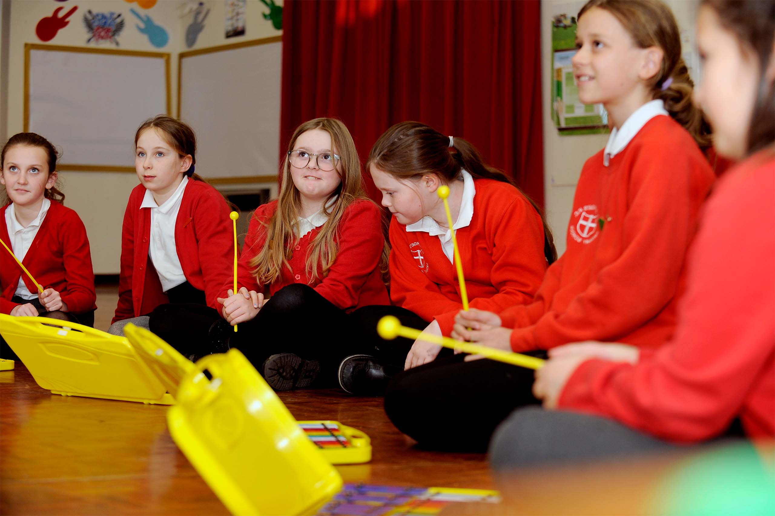 A group of focused schoolchildren in red uniforms are seated on the floor, holding yellow mallets, ready to play the colorful xylophones in front of them, fostering a sense of anticipation and musical collaboration in the classroom.