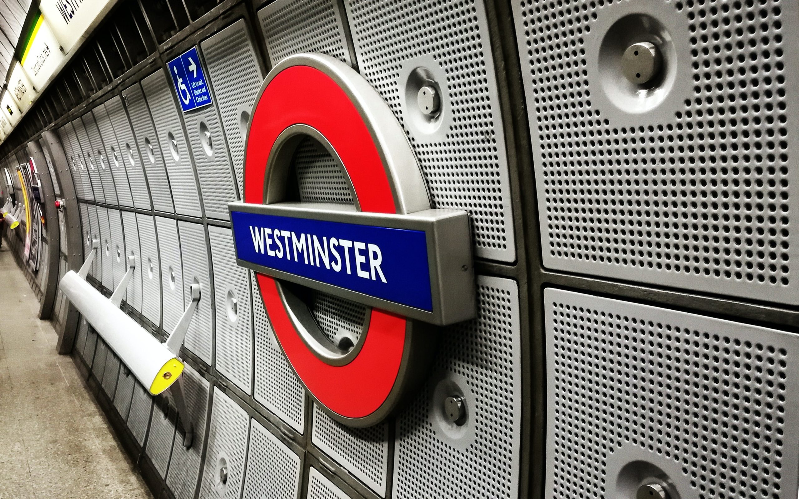 The photo shows the iconic roundel sign for Westminster station on the London Underground. The sign, with its bold red circle and blue bar bearing the station name in white lettering, is mounted on a textured, perforated metal wall characteristic of many tube stations. The perspective of the image leads down the platform, with the curvature of the tunnel visible in the distance, and a few passengers are present further along the platform.