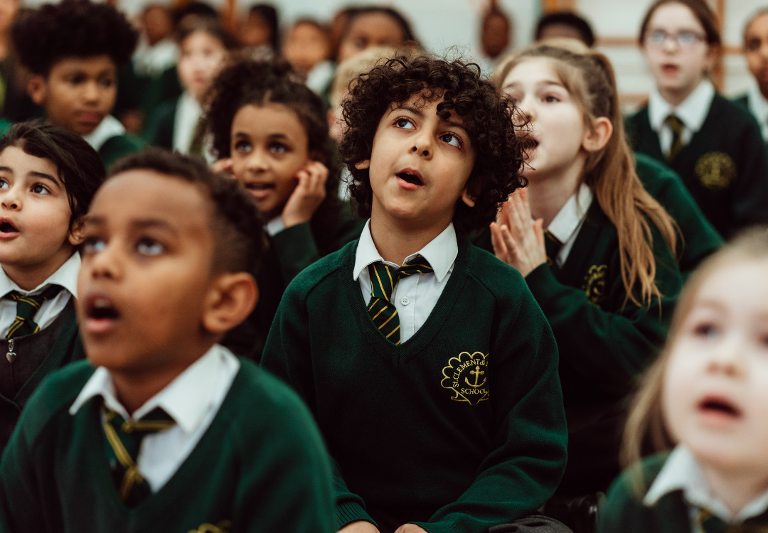 A group of schoolchildren, wearing green sweaters with school crests, are captured in a moment of wonder and anticipation. Their expressions range from awe to curiosity, with some looking upwards, mouths agape, and eyes wide, fully immersed in the experience unfolding before them. The atmosphere suggests a collective engagement, likely in response to an educational or entertaining event, highlighting the children's captivation and eagerness to learn.