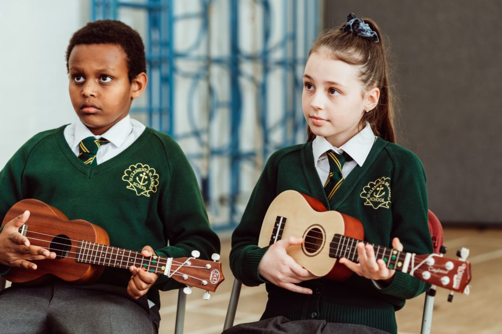 Two schoolchildren in green sweaters with embroidered school crests are seated and attentively holding ukuleles, seemingly ready for a music lesson. The boy on the left looks slightly away with a thoughtful expression, while the girl on the right gazes forward, her hands poised to play her instrument. Their focus suggests a moment of learning and concentration in a musical setting.