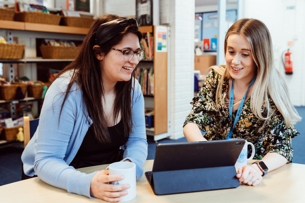 Two women are sharing a joyful moment at a table, with one holding a mug and the other interacting with a tablet. They are engaged in a friendly discussion, surrounded by a classroom environment filled with educational materials. Their smiles and relaxed postures suggest a warm, collaborative atmosphere.