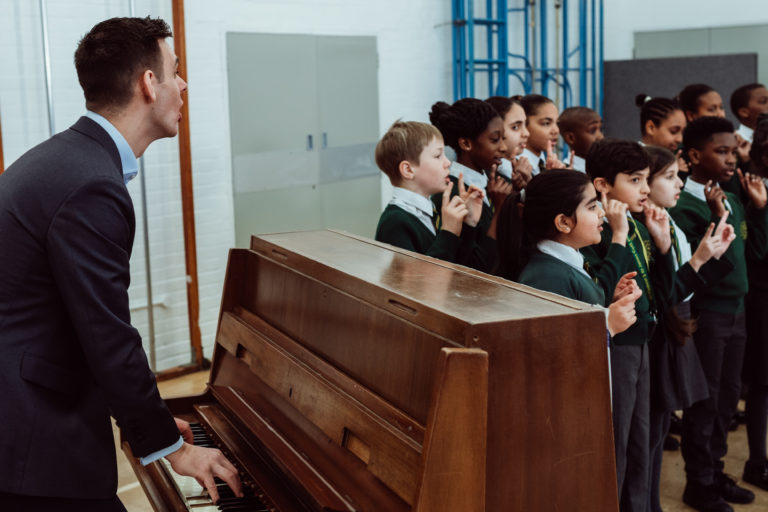 A group of schoolchildren, clad in green uniforms, are singing enthusiastically, accompanied by a man playing a piano. The children appear focused and joyful, participating in a harmonious choir session that fills the room with a sense of musical collaboration and learning.