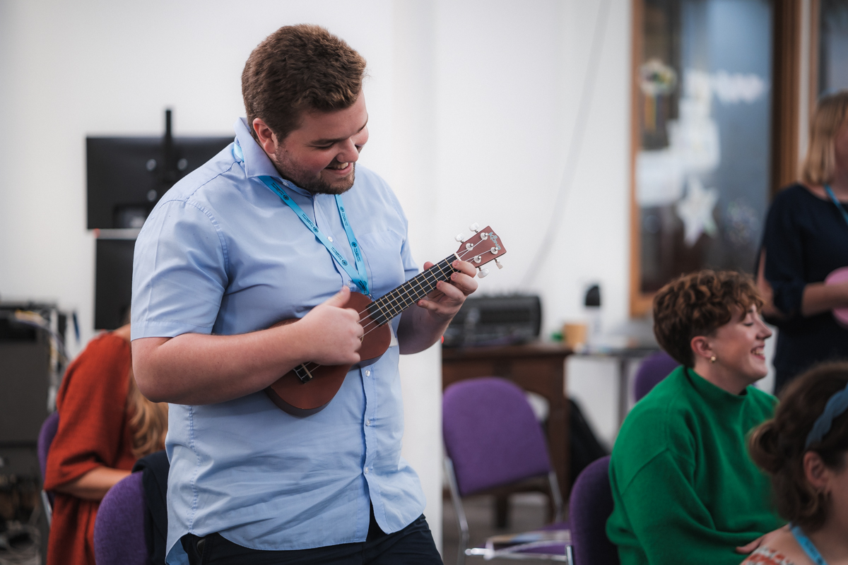 A man is playing a ukulele with focus and enjoyment, standing in a classroom setting where other participants are visible in the background, seemingly engaged in the music and the learning experience. The atmosphere suggests a collaborative and joyful educational environment.