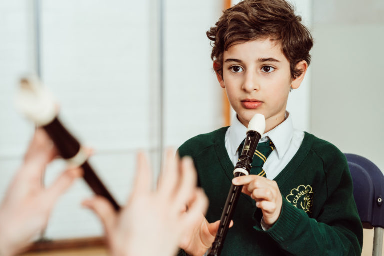 A young student in a green school sweater attentively holds a recorder, preparing to play, while a teacher's hands are seen in the foreground, guiding the child with a mallet, suggesting a musical instruction setting that fosters learning and creativity.