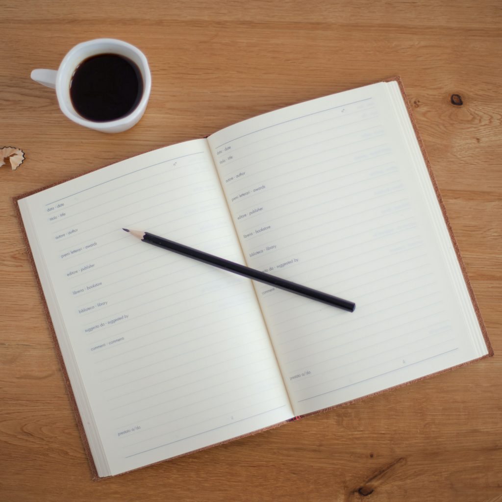 The image shows an open planner on a wooden surface with a black pencil resting on it. To the left of the planner, there's a white cup filled with coffee. The planner has printed dates and lines for notes, but no writing, suggesting a fresh start or planning ahead. The scene conveys a sense of organization and productivity, with a hint of a cozy atmosphere provided by the warm tone of the wood and the presence of the coffee.