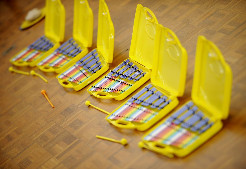 A series of yellow cases opened to reveal colorful xylophones, each paired with mallets, are neatly arranged on a wooden floor, suggesting an inviting and organized setting for a music class or ensemble practice.