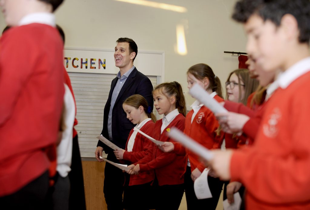 A group of focused schoolchildren in red uniforms, holding sheets of music, are engaged in a singing session led by an enthusiastic instructor. The instructor, standing among the students with a beaming smile, guides the choir with visible passion, creating an atmosphere of joyful learning and musical collaboration.