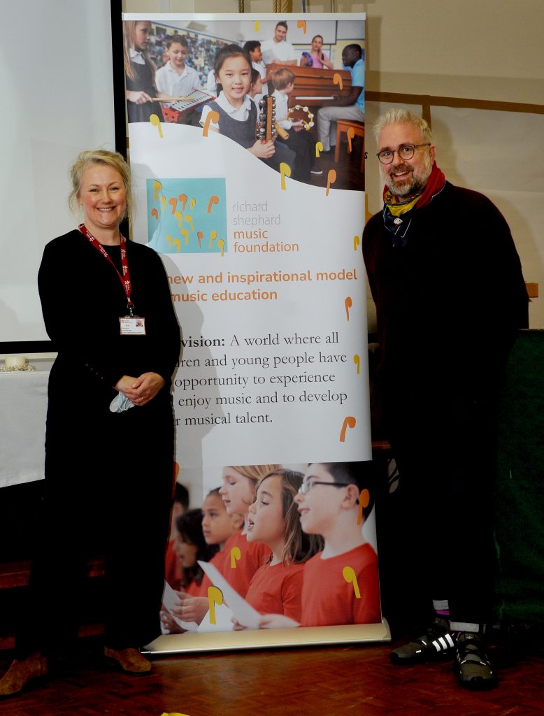A man and a woman stand smiling on either side of a promotional banner for the Richard Shephard Music Foundation, which displays images of children engaged in music education and text outlining the foundation's vision for accessible music education. The atmosphere suggests a supportive and proud endorsement of the foundation's mission to inspire and develop musical talent in young people.