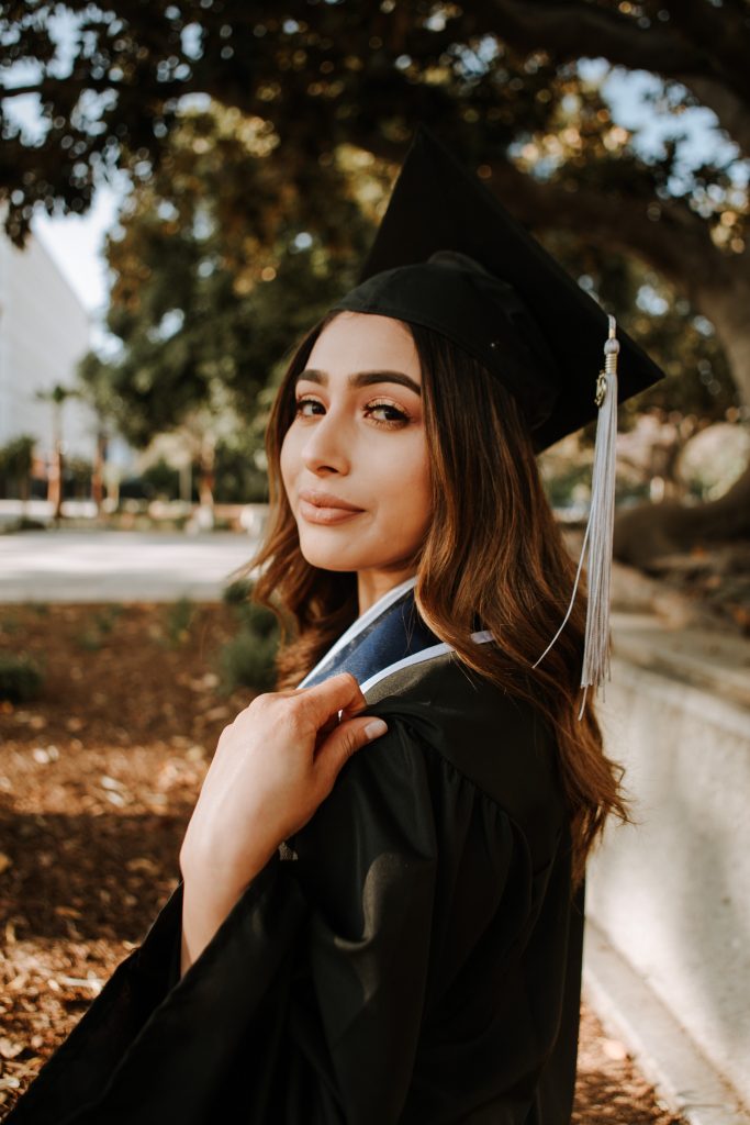 A confident young woman in graduation attire stands outdoors, her gaze slightly off-camera, exuding a sense of accomplishment and optimism. The tassel of her mortarboard gently sways, complementing her poised stance as she adjusts her graduation stole, symbolizing her academic achievements. Trees and soft sunlight create a serene backdrop, highlighting this momentous occasion in her educational journey.