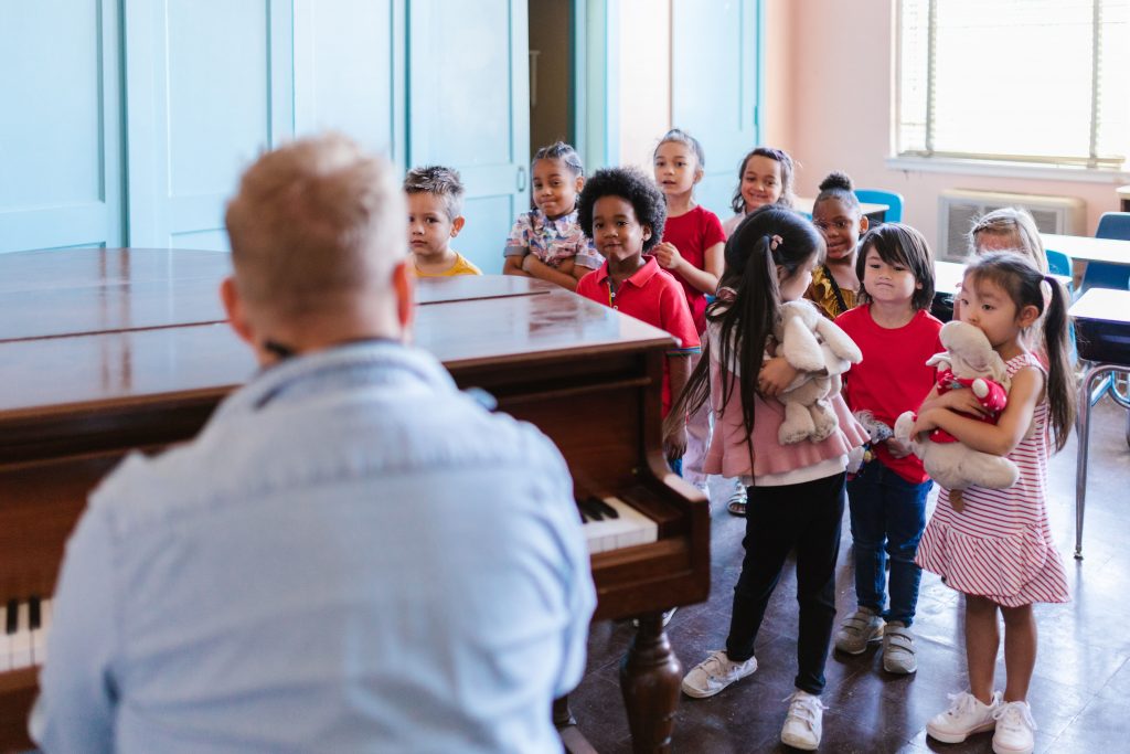 A group of young, diverse students stands attentively in a classroom, their faces expressing curiosity and anticipation as they watch an instructor play the piano, hinting at a musical lesson that engages and inspires. Some children hold stuffed animals, adding a touch of comfort and playfulness to the learning environment.