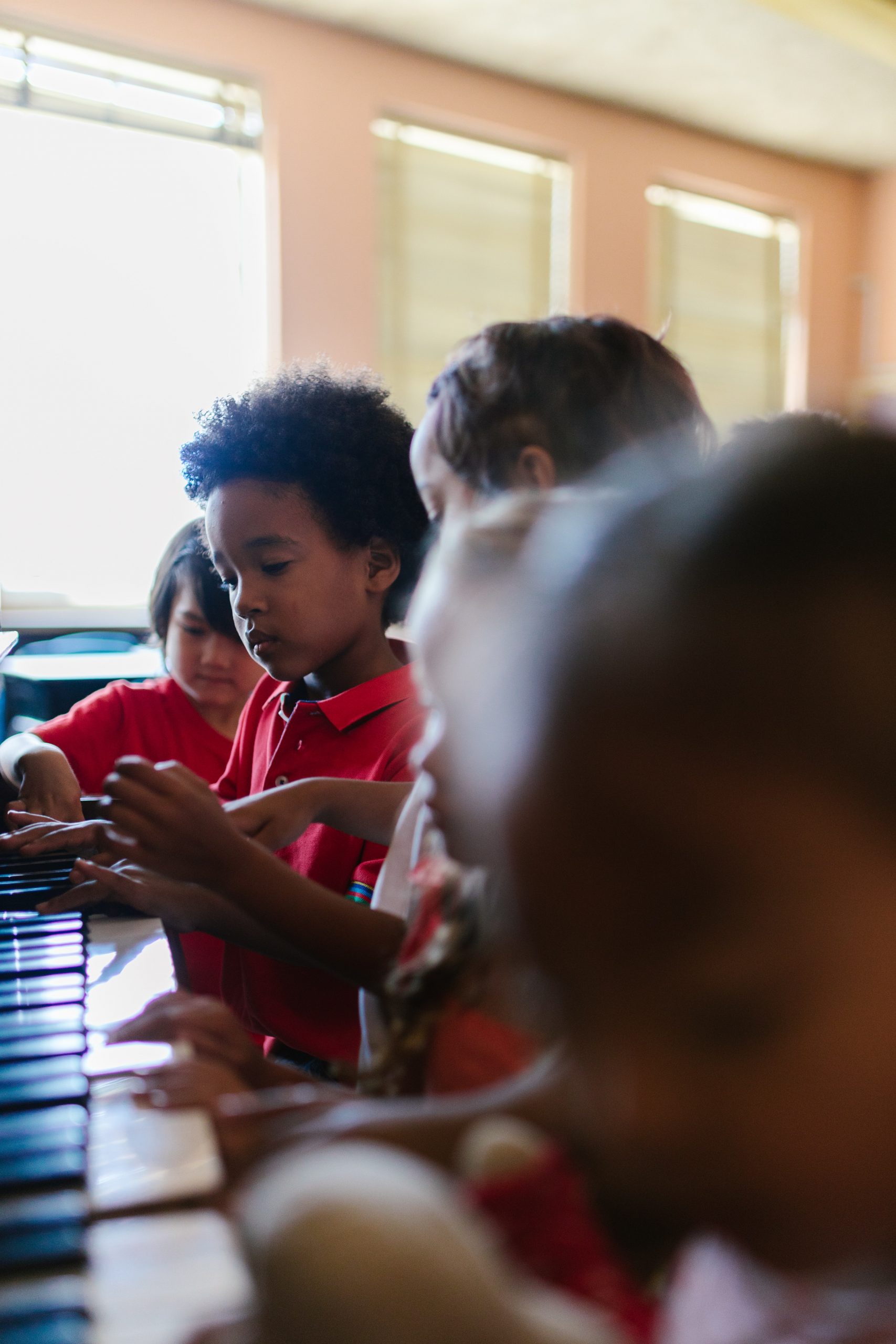 A focused child in a red shirt is playing a piano, surrounded by classmates in a sunlit classroom, capturing a moment of musical exploration and peer learning.