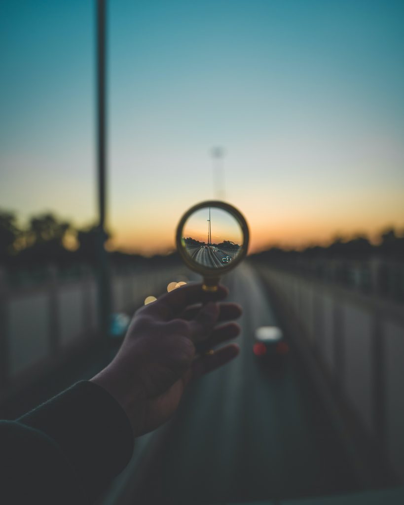 The photo captures a hand holding a crystal ball that reflects and inverts the image of a road stretching into the distance during sunset. The background is out of focus, with the bridge, sky, and ambient lights softly blurred, while the scene within the crystal ball is sharp and clear. The setting sun casts a warm glow, enhancing the tranquil and somewhat mystical atmosphere of the moment.