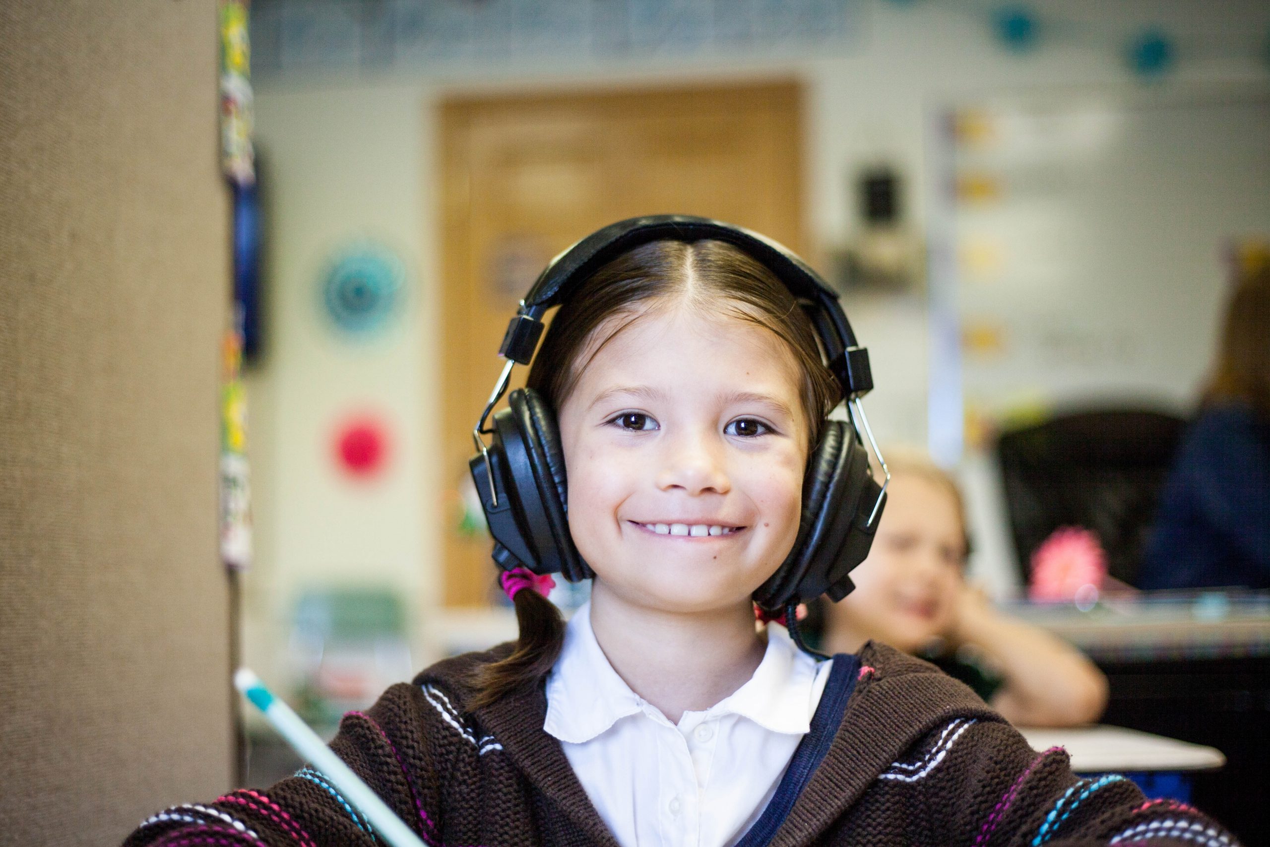 A young student with a beaming smile is wearing large headphones, sitting in a classroom setting. Her bright eyes and cheerful expression suggest engagement and enjoyment, possibly listening to music or engaging in an interactive learning activity. The colorful classroom environment with artwork in the background adds to the lively and positive educational atmosphere.