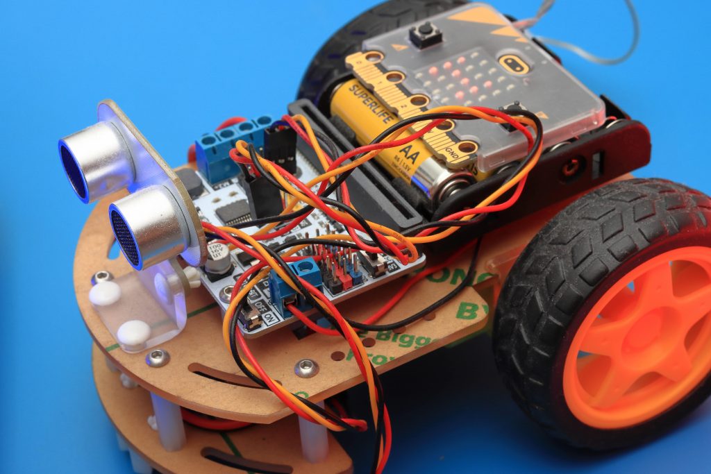 The image shows a close-up of a DIY robotic car kit with a blue background. The robot has a wooden chassis and is equipped with various electronic components including a microcontroller board, sensors, a battery pack, and a tangle of colorful wires. The robot also features two large black wheels on one side and a smaller orange wheel visible on the other, suggesting it's a three-wheeled vehicle. The ultrasonic sensors at the front resemble a pair of eyes, giving the robot a somewhat anthropomorphic appearance.