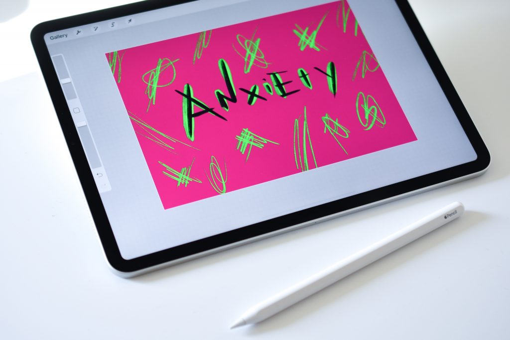 The image shows a digital tablet with a bright pink canvas displayed on its screen, featuring the word "Anxiety" written in black amidst various green scribbles and marks. A stylus, presumably used for drawing or writing on the tablet, lies in the foreground. The overall scene suggests digital artwork or the expression of feelings through a graphic design app.