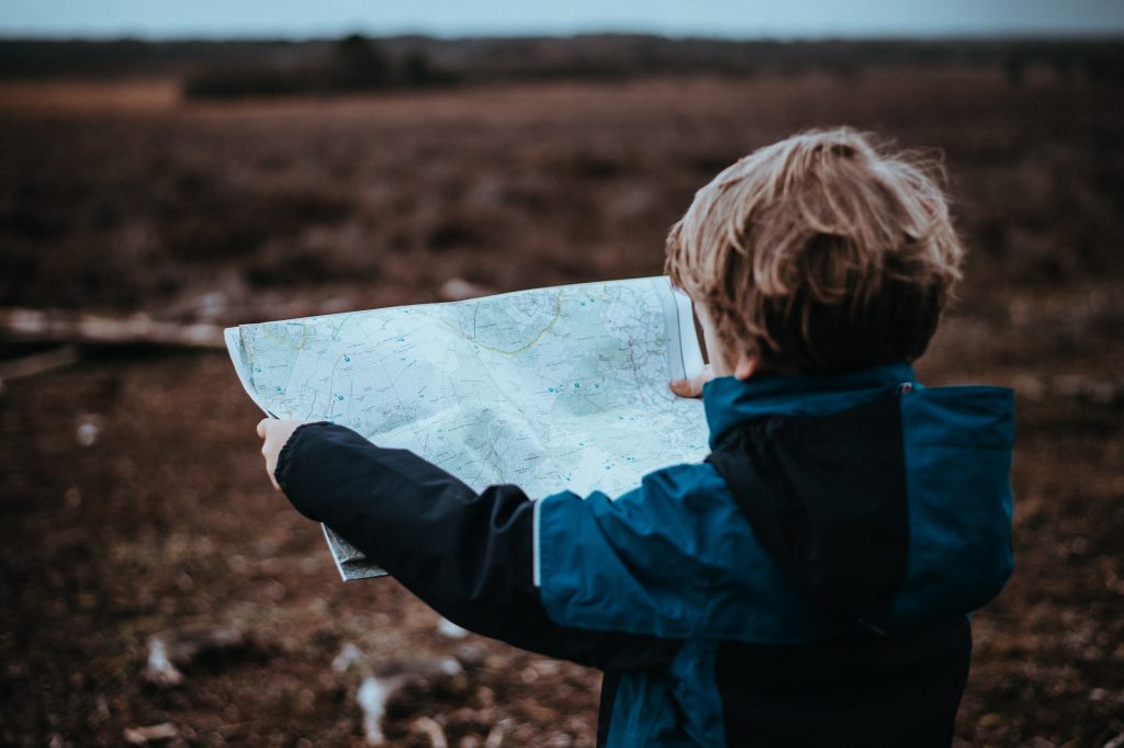 A young child with tousled hair is standing in a barren field, holding a large, unfolded map with both hands, seemingly engrossed in navigating the landscape. The child's back is to the camera, and they are wearing a blue jacket, suggesting an adventurous spirit on a cool day. The focus on the map and the child's engagement with it evoke a sense of exploration and discovery.