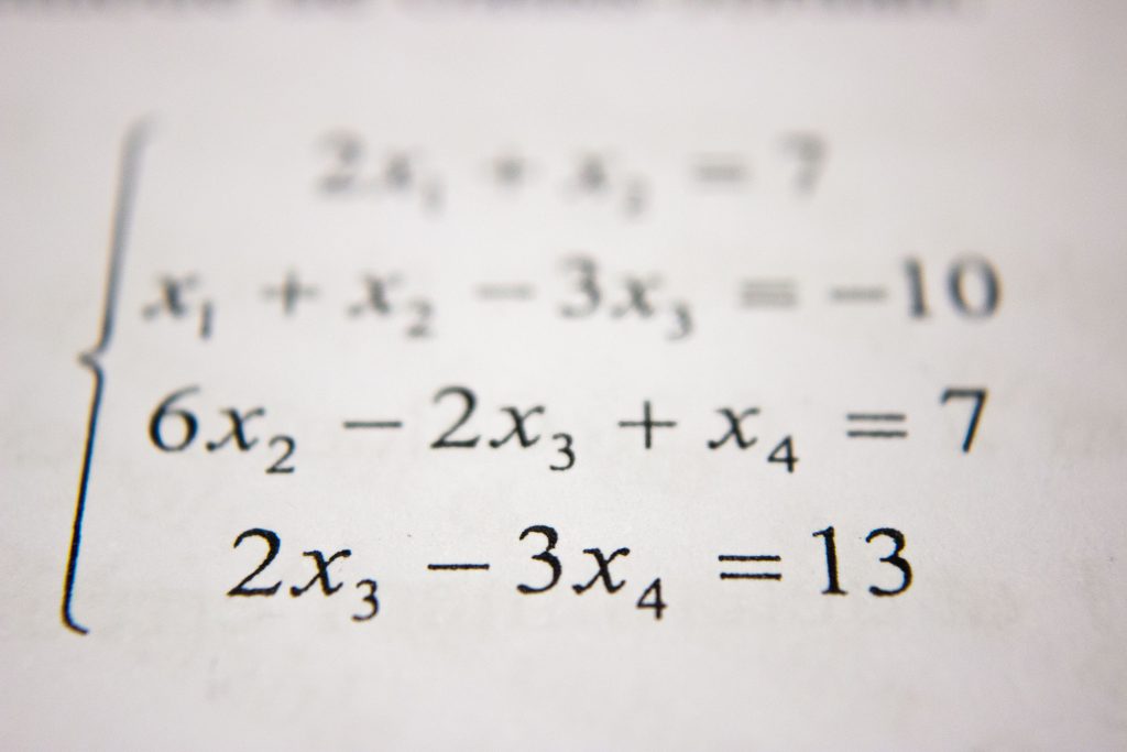 The image shows a close-up of a printed page with a set of mathematical equations, specifically a system of linear equations with variables x1, x2, x3, and x4. The focus is shallow, with the foreground and background blurred, highlighting the middle equation which reads "6x2 - 2x3 + x4 = 7". The equations are part of an algebraic problem, possibly for an educational purpose such as homework or a textbook example.