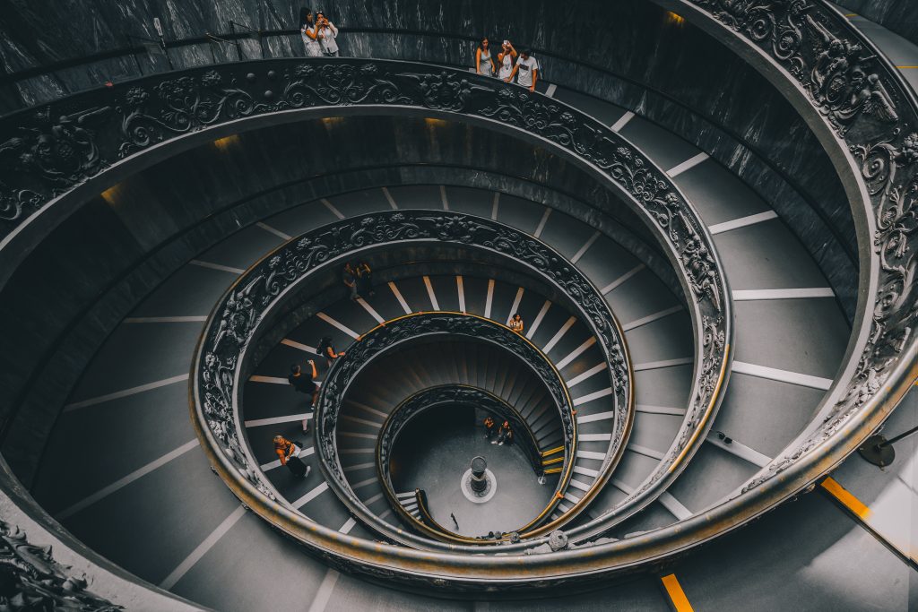 The image shows the iconic double helix spiral staircase, often associated with the Vatican Museums. This architectural marvel features two intertwined staircases that allow for separate paths of ascent and descent. The ornate balustrades and the smooth, sweeping curves of the steps create an elegant visual effect. Visitors can be seen capturing the moment on their cameras, admiring the staircase from various levels. The overall atmosphere is one of awe and appreciation for this unique and beautiful structure.