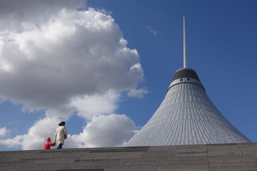 The image shows a person and a child ascending a flight of outdoor steps, with the child wearing a bright red coat. In the background, a large conical building with a distinctive spire reaches into a sky dotted with fluffy clouds. The building's surface appears to be made of ribbed metal or similar material, giving it a textured appearance. The scene is composed with a sense of upward movement and aspiration, emphasized by the grandeur of the architecture and the expansive sky.