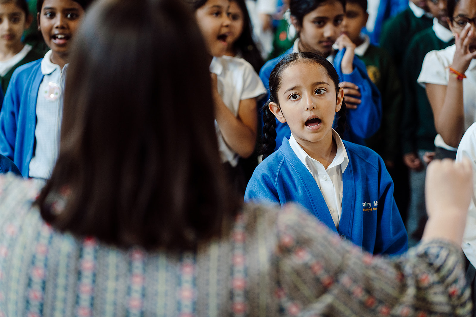 A group of enthusiastic schoolchildren are singing in a classroom, with a focus on a young girl in a blue sweater and white shirt, who is looking intently at the teacher leading the activity. The children's expressions convey engagement and enjoyment as they participate in the musical learning experience.