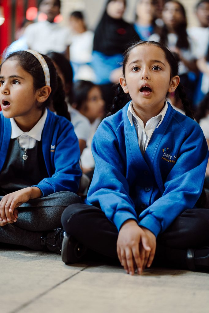 Two young students, wearing blue school sweaters, are seated on the floor with a look of concentration and wonder. The girl in the foreground is singing with an open mouth, suggesting active participation in a classroom activity, while her peers in the blurred background appear to be equally engaged. The atmosphere conveys a sense of immersion and enjoyment in the learning experience.