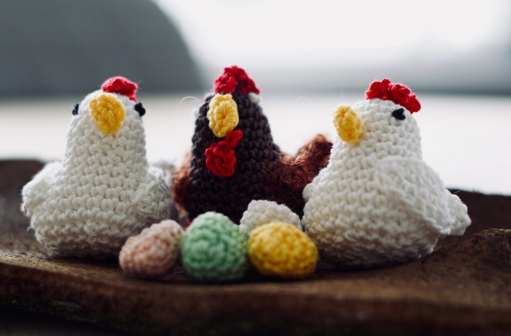 Three crocheted chickens with red combs and beaks are positioned on a wooden surface, accompanied by several colorful crocheted eggs. The craftsmanship gives the chickens a charming and whimsical appearance, evoking a cozy, handmade aesthetic.