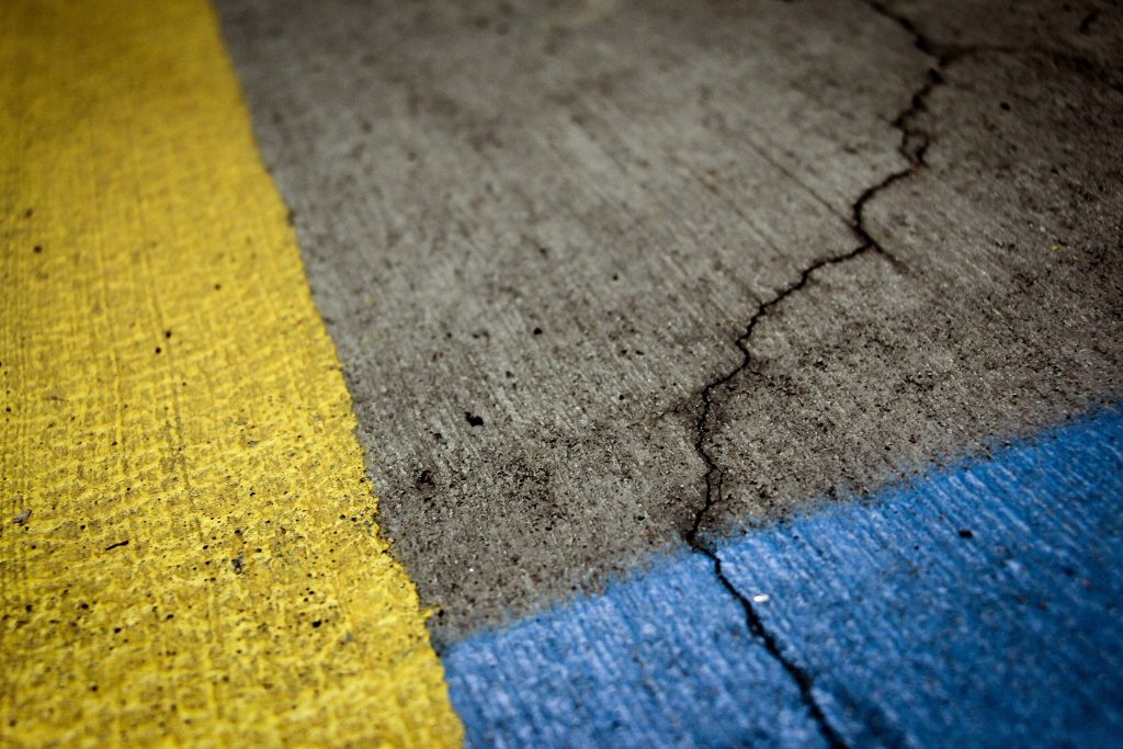 The image shows a close-up of a concrete surface with painted lines in yellow and blue, indicating a parking or no-parking zone. There's a visible crack running through the concrete, adding texture and a sense of wear to the scene. The perspective is angled, focusing on the boundary where the painted lines meet the grey concrete.